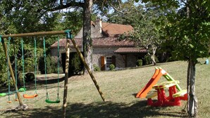 A playground with swings and slides, a stone house with a tiled roof, and a grassy area with trees.