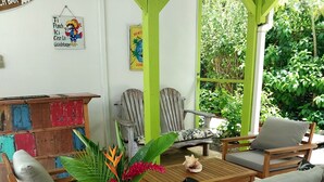 Een houten zithoek buiten met een tafel en stoelen, omringd door groen en een stenen vloer.