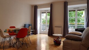 A living room with a dining area, a sofa, a TV, and a window with curtains. - L'EUROSEBASTOPOL - Crossing apartment  (Strasbourg)