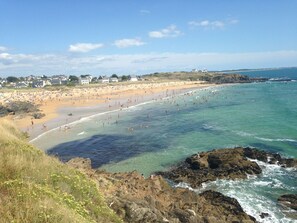 Escena de playa costera con aguas transparentes, costa rocosa y edificios al fondo.
