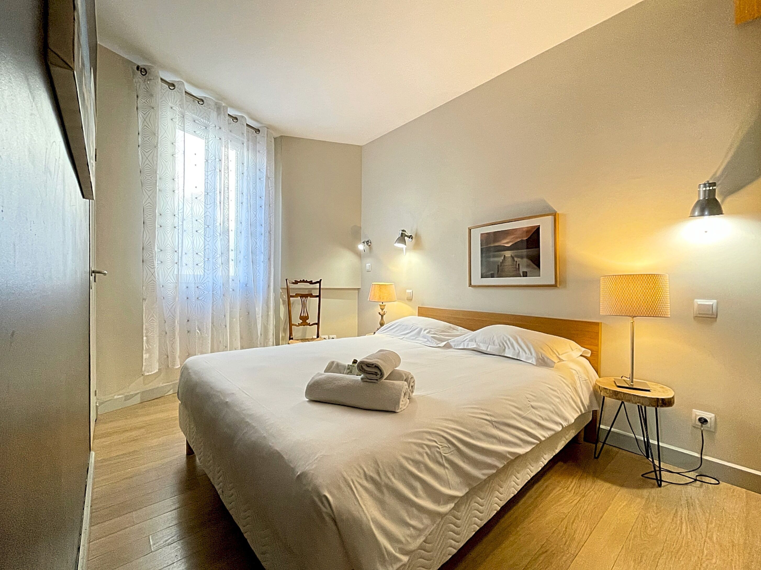 Hotellihuone, jossa on suuri sänky, kaksi yöpöytävalaisinta, seinällä kehystetty taulu ja pieni pyöreä pöytä, jossa on valaisin.