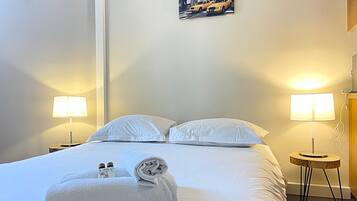 A hotel room with a neatly made bed, two bedside lamps, a cityscape painting on the wall, and a small wooden side table with a lamp.