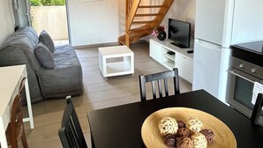 A modern living space with a wooden staircase, a sofa, a dining table with a bowl of fruit, a kitchen area with a refrigerator and oven, and a TV on a white cabinet. - T2 apartment, 40 m2 350m from Lecques beach, quiet. Air conditioning. Private parking (Saint Cyr sur Mer)