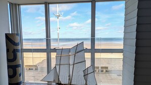 Une maquette de bateau est exposée sur un présentoir dans une pièce dotée de grandes fenêtres donnant sur une plage.
