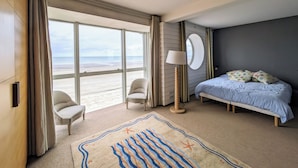 Une chambre d’hôtel avec une grande fenêtre, un lit, deux chaises, une lampe sur pied et un tapis à motifs de plage.