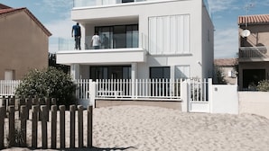 A modern two-story house with a balcony, a white fence, and a sandy beach in the foreground. - Superb new apartment "feet in the water" exceptional view on the beach (Mauguio)