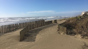 A sandy beach with a wooden fence leading to the ocean, a clear sky, and buildings in the background. - Superb new apartment "feet in the water" exceptional view on the beach (Mauguio)
