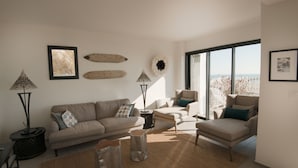 A modern living room with a sofa, armchairs, and a coffee table. There are decorative wall hangings and a large window with a view. - Superb new apartment "feet in the water" exceptional view on the beach (Mauguio)