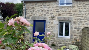 A stone house with a blue door and window, a garden with pink flowers, and a small table with a chair.