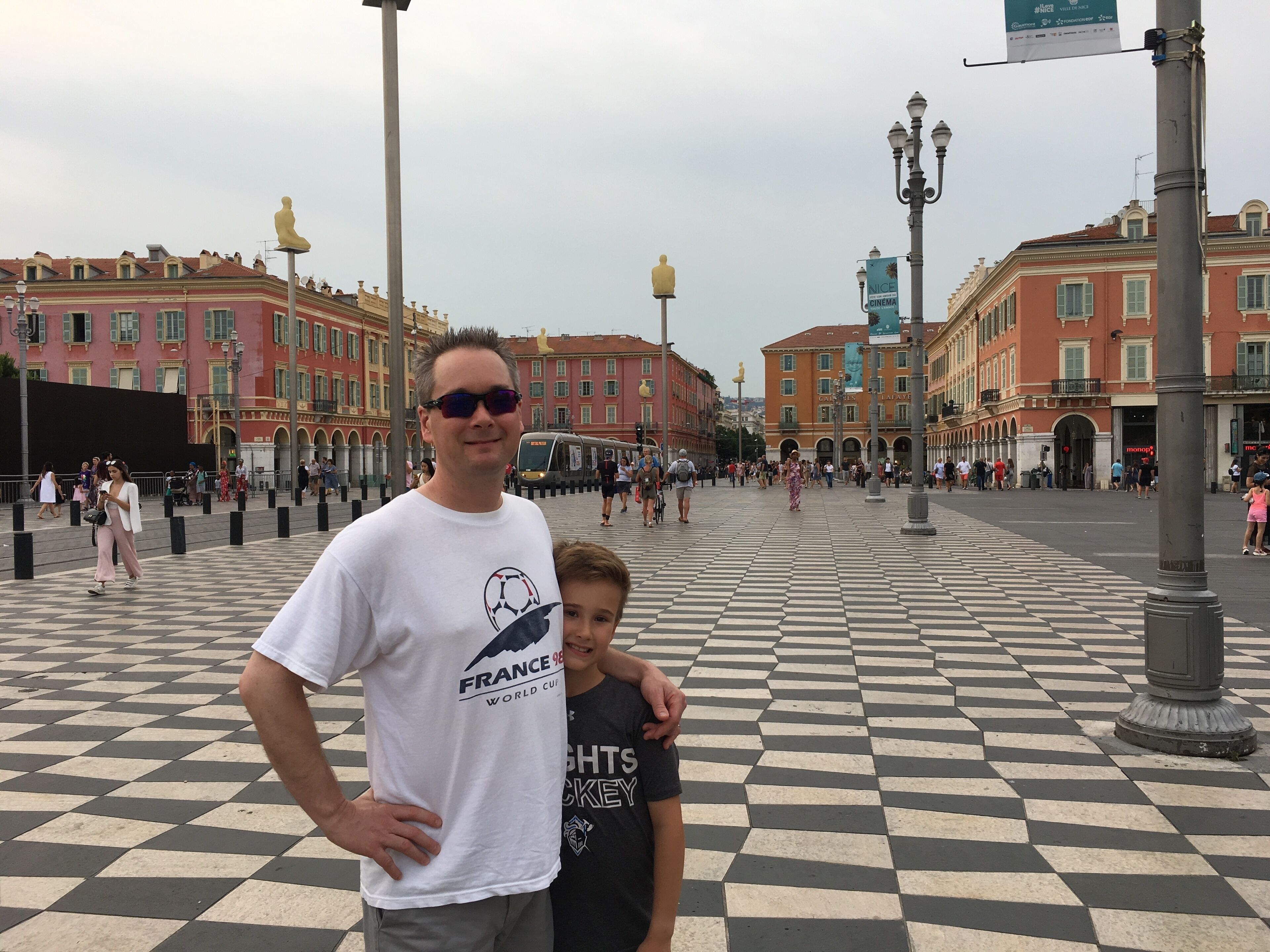 A man and a child standing in a public square with a checkered floor, surrounded by historic buildings.