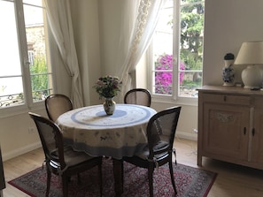Uma área de refeições com uma mesa redonda, cadeiras, um vaso com flores, um abajur e vista para o verde pela janela.