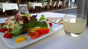 Une table avec une assiette de salade et un verre d’une boisson claire, probablement du vin, dans un espace de restauration en plein air, avec des chaises et des tables en osier.