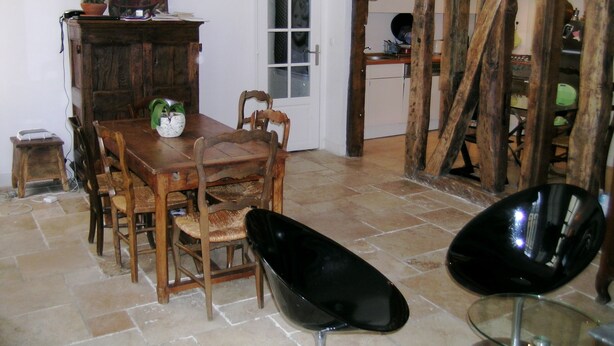 A dining area with a wooden table, chairs, and a glass-top table. There is a cabinet with a plant on top and a door leading to another room.