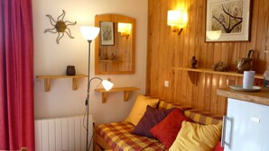 A cozy room with a wooden bed, a small table, a chair, a refrigerator, and a shelf with decorative items.