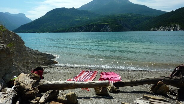 A rocky beach with a wooden log barrier, a red and white striped blanket, and a person in red clothing.