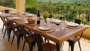 Une table à manger dressée pour six personnes, avec des chaises, une bouteille de vin et un vase de fleurs, sur un balcon offrant une vue sur un paysage verdoyant.