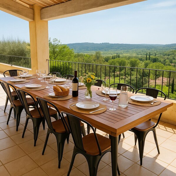 A dining table set for six with chairs, a bottle of wine, and a vase of flowers on a balcony with a view of a green landscape.