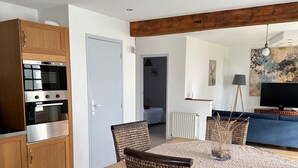 Une cuisine moderne avec des meubles en bois et un four encastré, un coin repas avec des chaises en osier et une table en bois, et un salon avec un canapé bleu et un tableau.