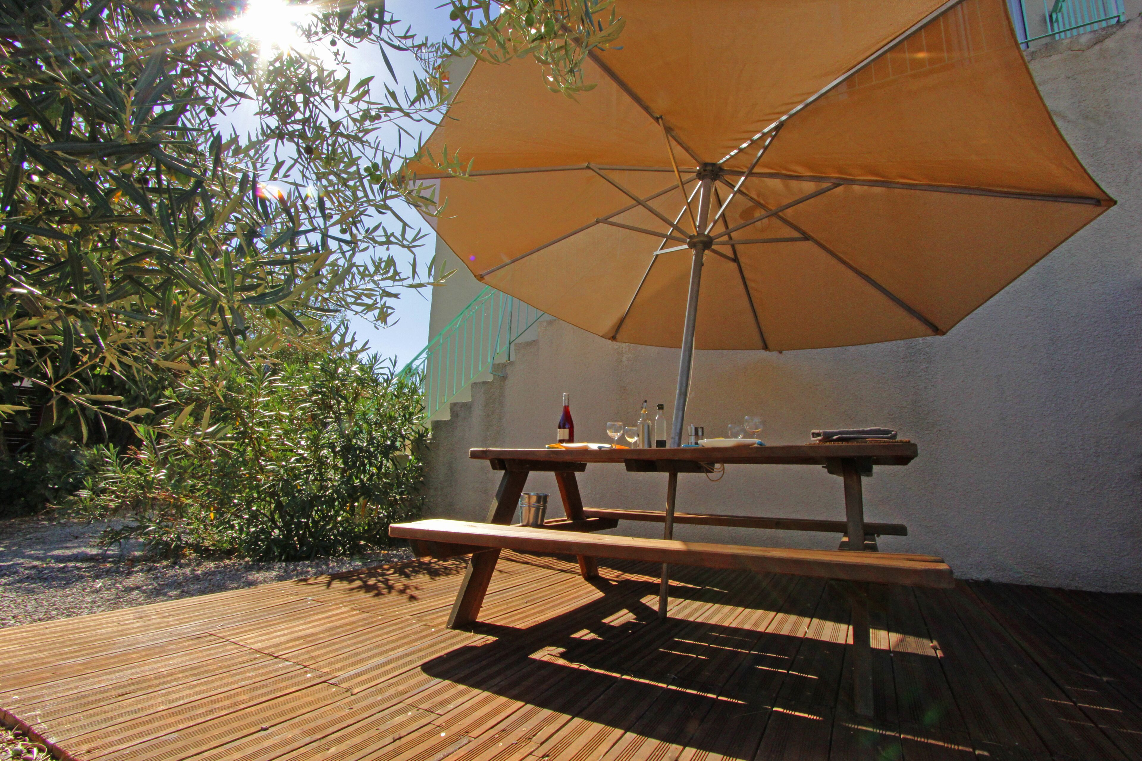 A wooden picnic table with an umbrella, surrounded by greenery and a white wall.