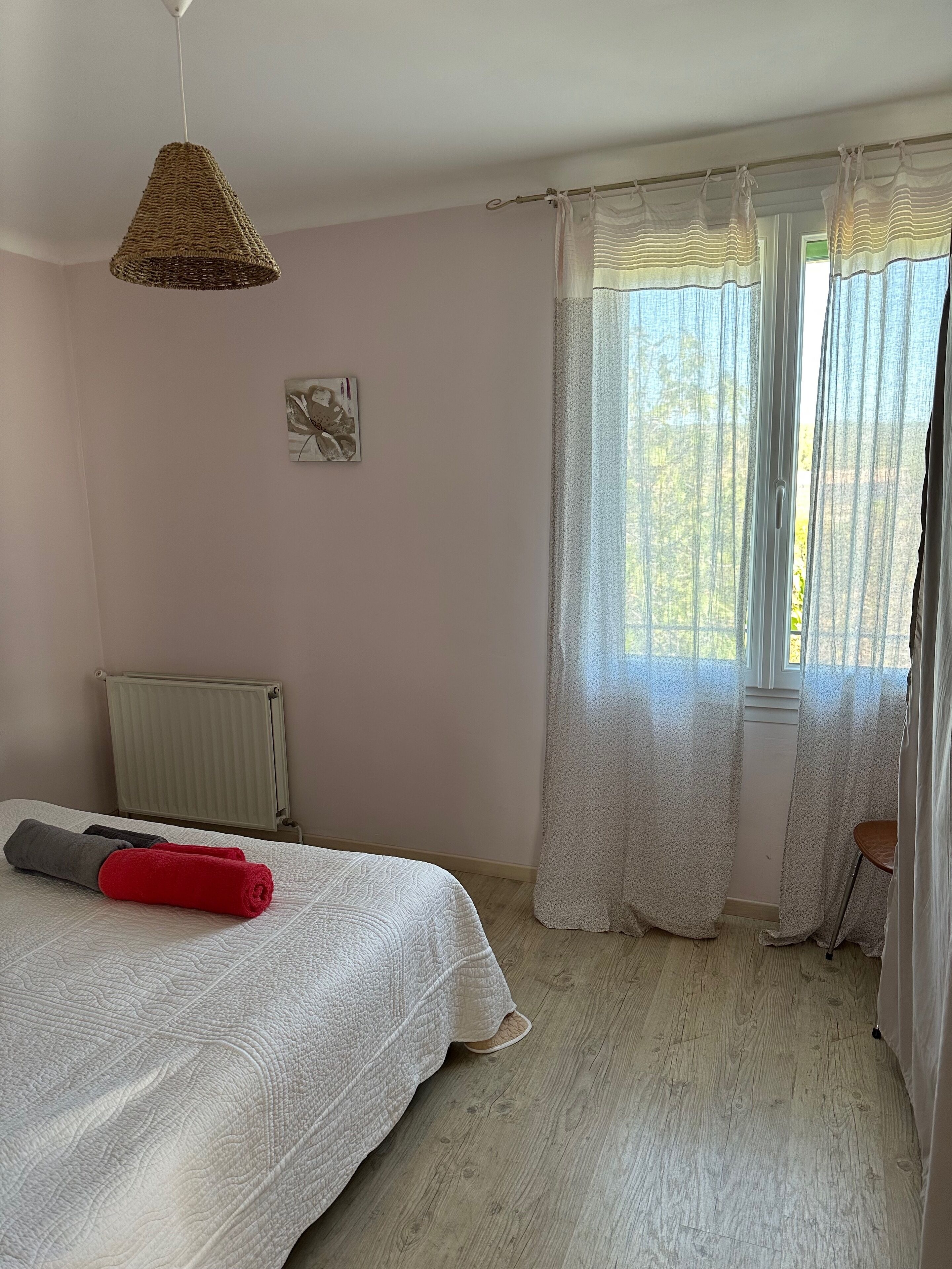 Une chambre à coucher avec un lit, un radiateur, un luminaire suspendu et une fenêtre ornée de rideaux légers.