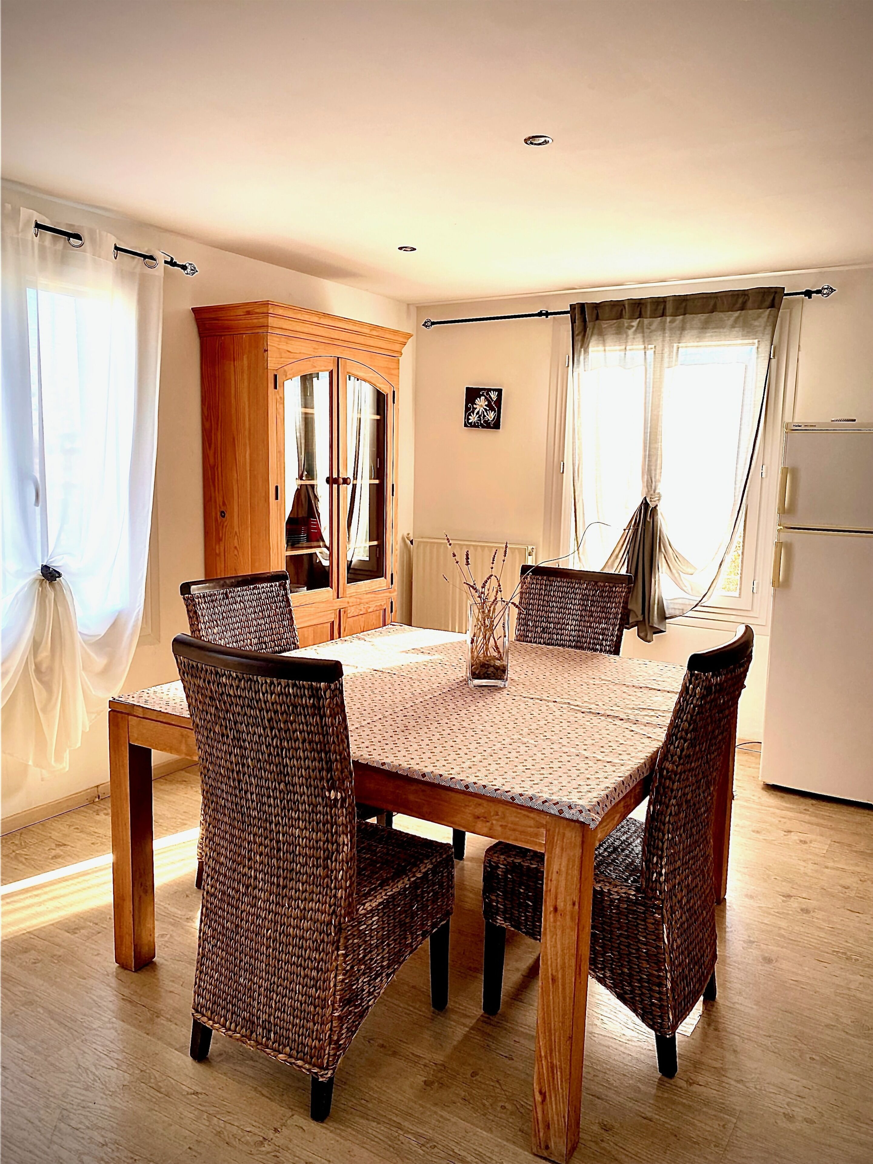 Un coin repas avec une table en bois et des chaises en osier, un buffet en bois et un réfrigérateur.