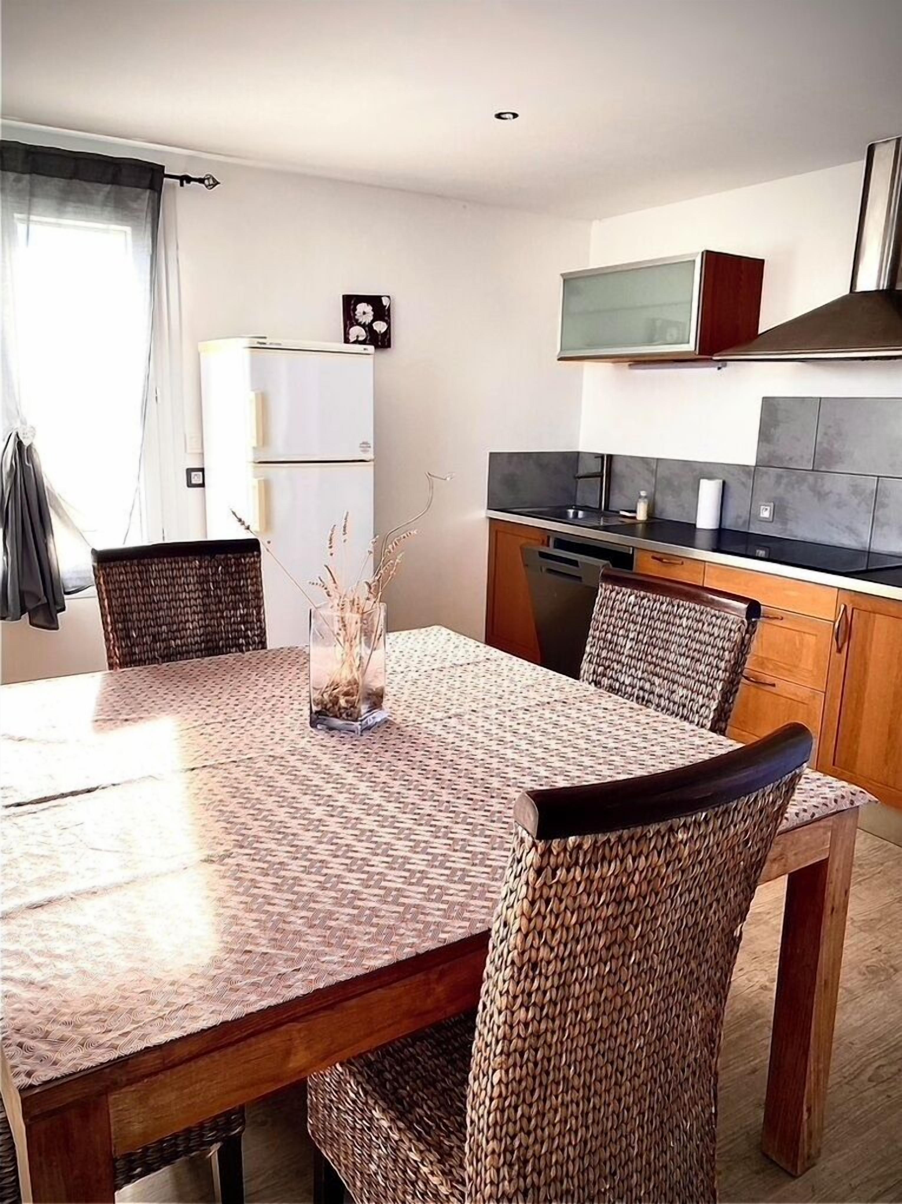 Uma sala de jantar com mesa e cadeiras de fibra, um frigorífico, uma cozinha com fogão e armários, e uma janela com cortinas.