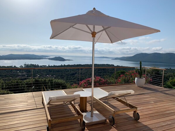 Terraza de madera con dos sillones reclinables y una sombrilla grande, con vista a un paisaje pintoresco de un lago y montañas lejanas.