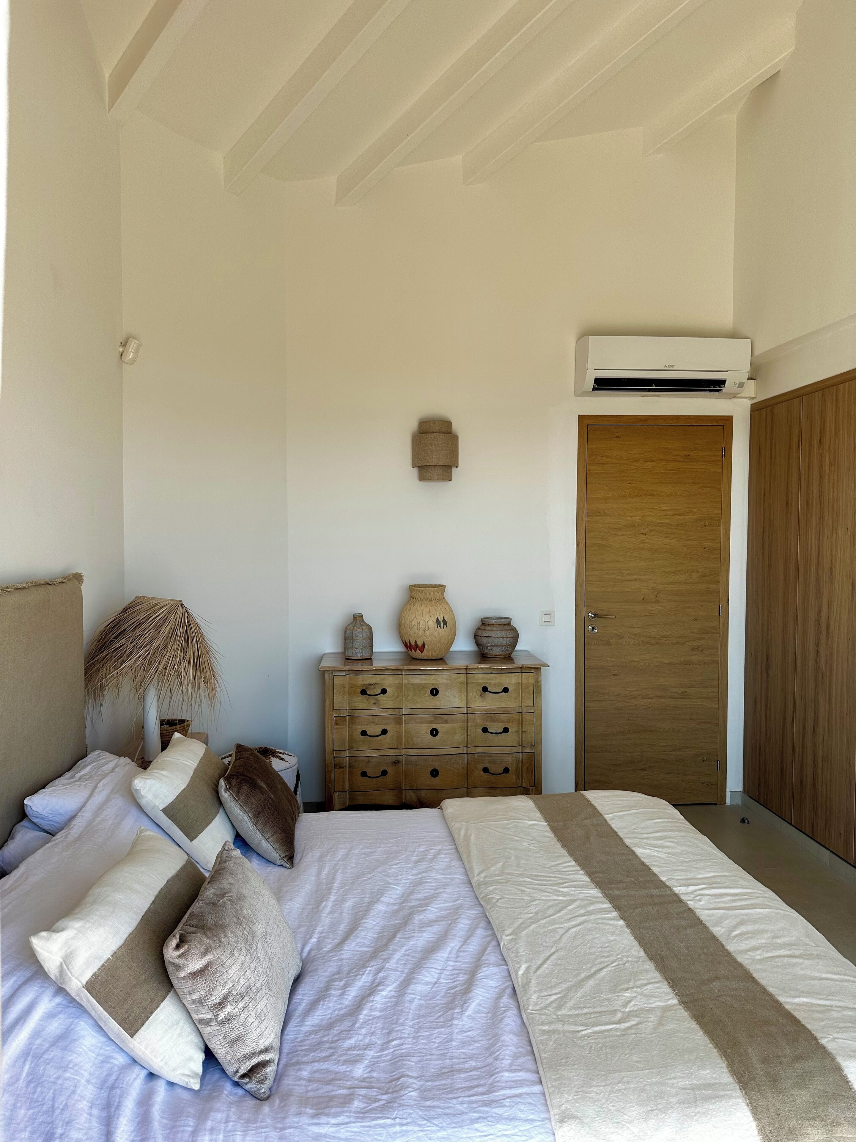 A bedroom with a bed, a wooden dresser, an air conditioning unit, and a wooden door.