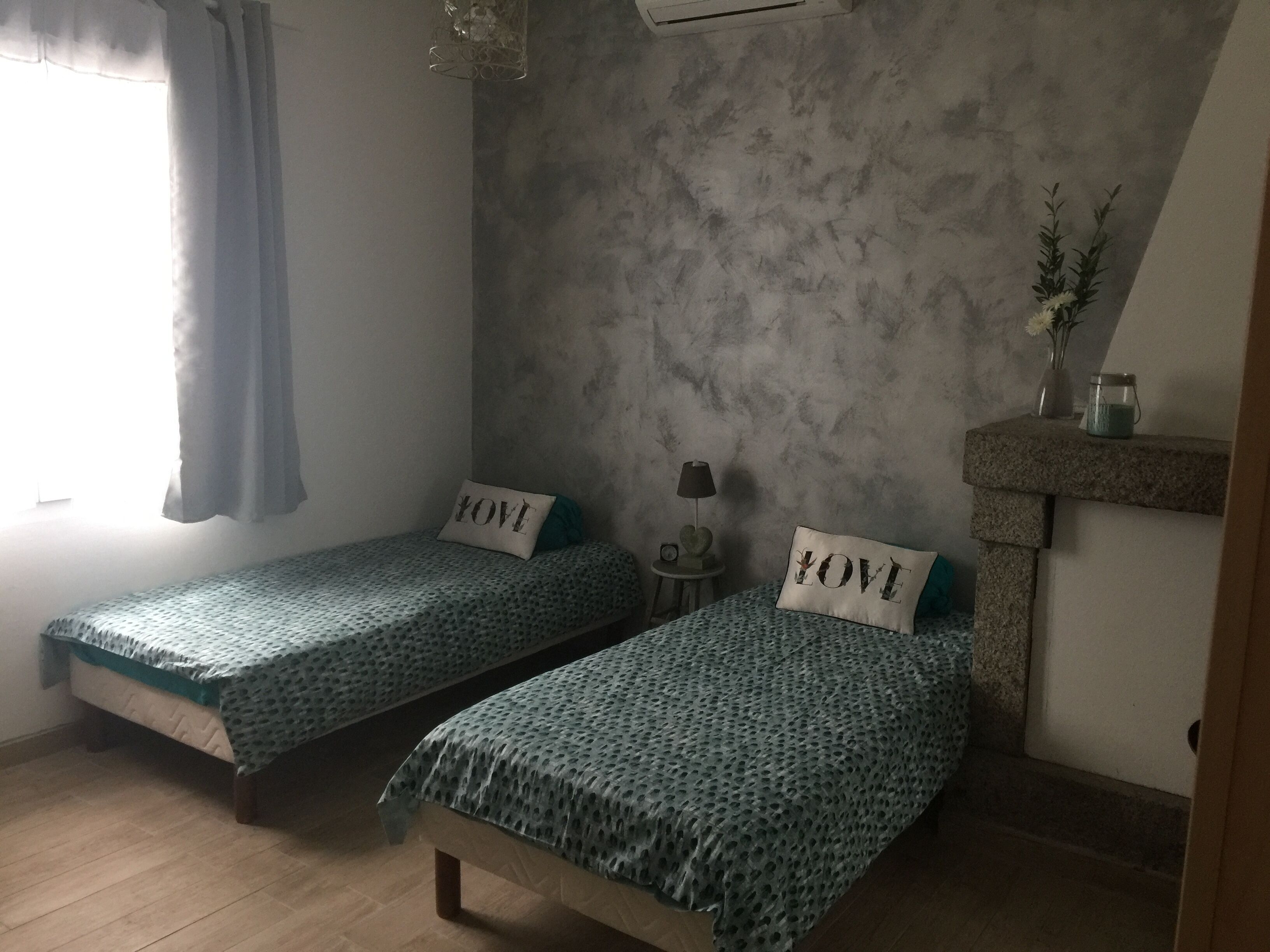 Un dormitorio con dos camas individuales, una mesita de noche con lámpara, un estante en la pared con un jarrón y una ventana con cortinas.