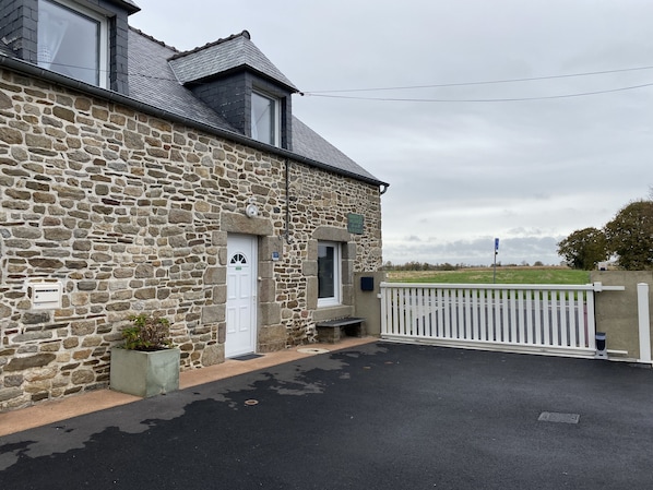 Un bâtiment en pierre avec une porte blanche, un petit porche et une allée d’accès fermée par un portail.