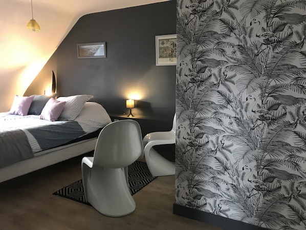 Et soveværelse med seng, to stole, et natbord, en lampe og væg med tropisk tapet.