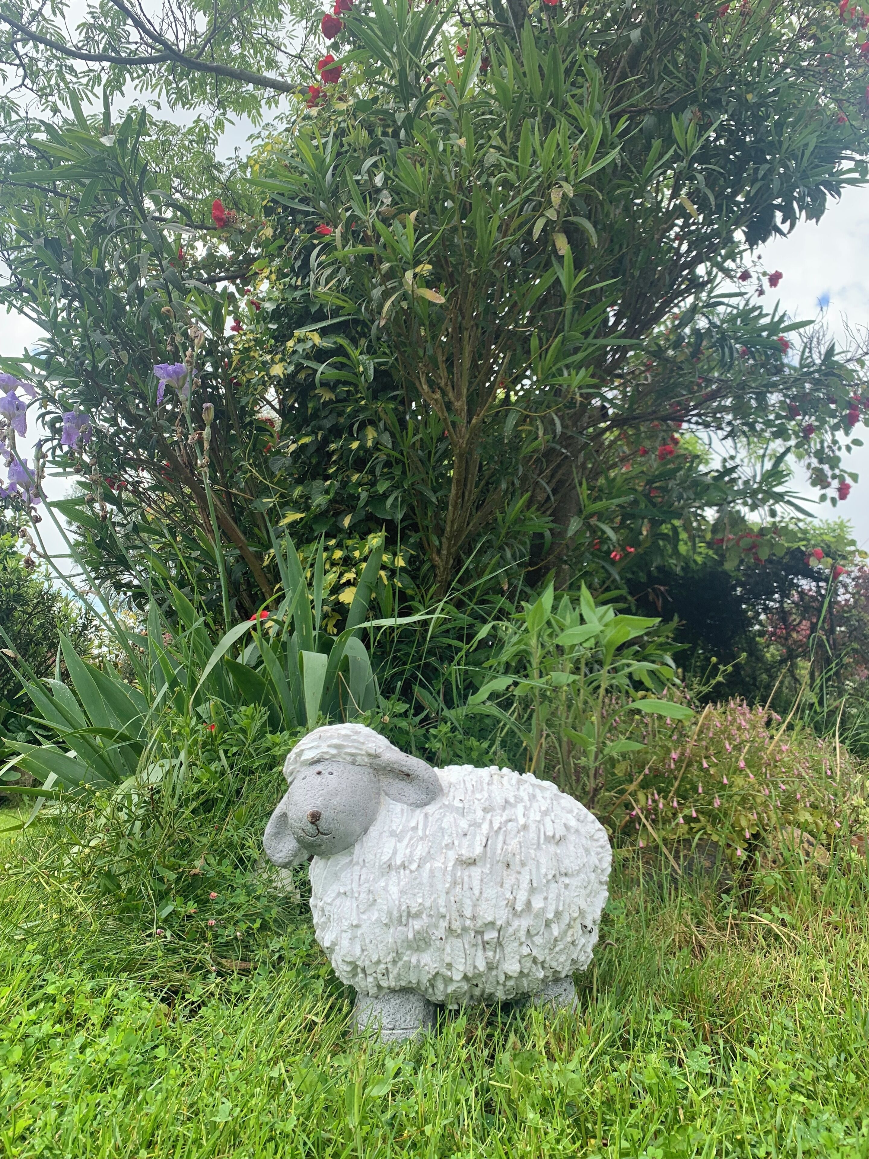 Un giardino con una scultura di una pecora bianca, circondato da vegetazione lussureggiante e piante in fiore.