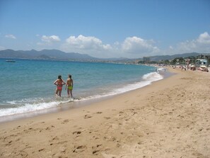 A beach with two people walking along the shore, a clear sky, and distant mountains.
