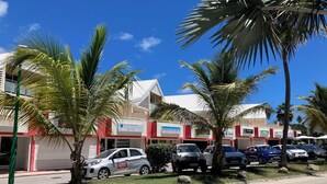 A street view with parked cars, palm trees, and buildings with signage. - BEAUTIFUL STUDIO NEAR THE BEACH IN THE HEART OF THE ORIENT BAY VILLAGE (Orient Baie)