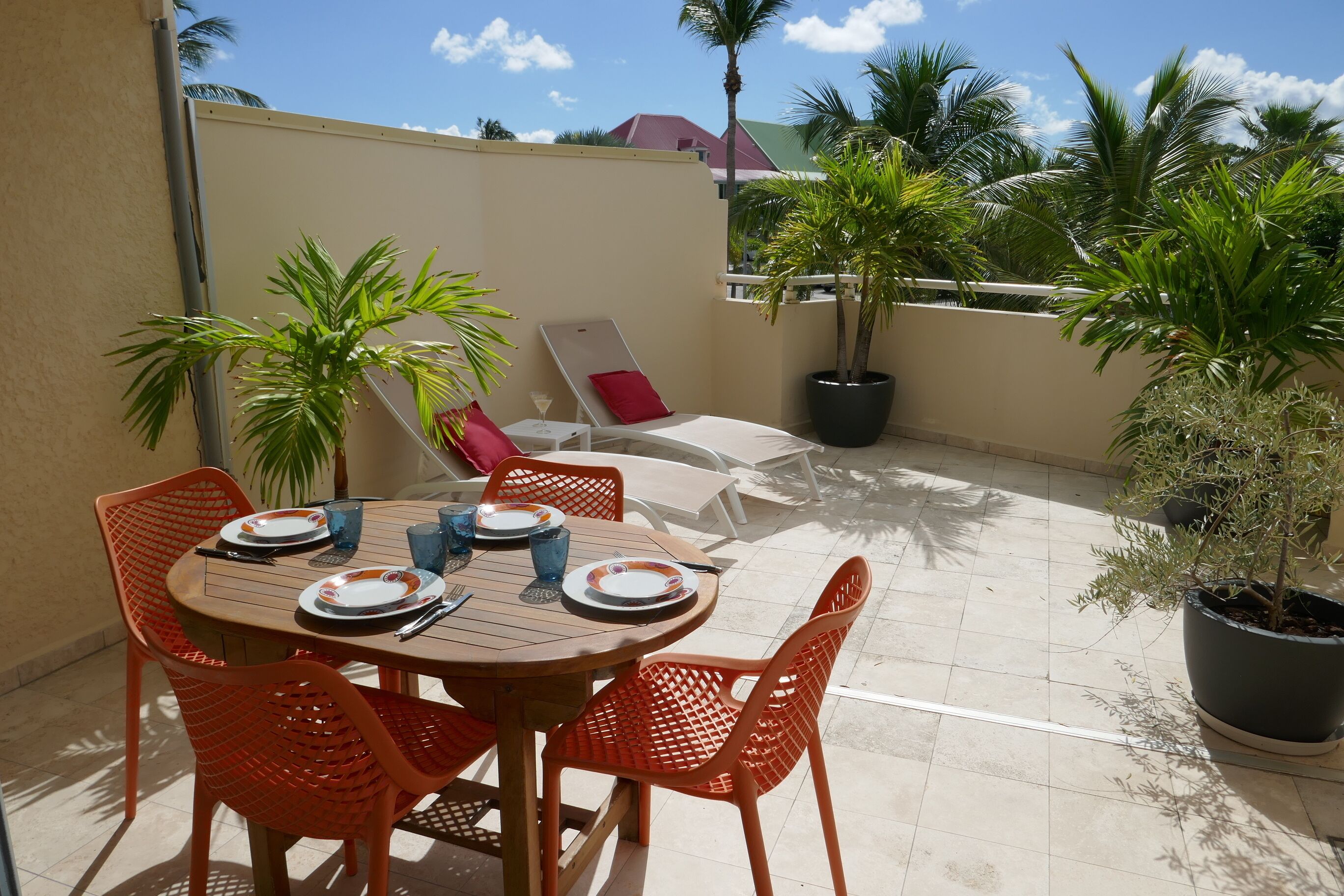 Eine Terrasse mit einem runden Tisch, orangefarbenen Stühlen und einem roten Kissen. Übertöpfe mit Pflanzen, im Hintergrund Palmen und ein Gebäude.