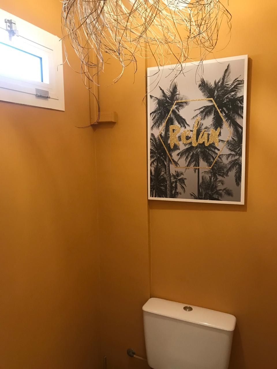 Un baño con un inodoro, un aplique de pared y un cuadro enmarcados con palmeras y la palabra 'Relax' escrita en la pared.