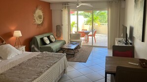 A hotel room with a bed, a green sofa, a coffee table, a desk with a computer, and a balcony with outdoor furniture. - BEAUTIFUL STUDIO NEAR THE BEACH IN THE HEART OF THE ORIENT BAY VILLAGE (Orient Baie)