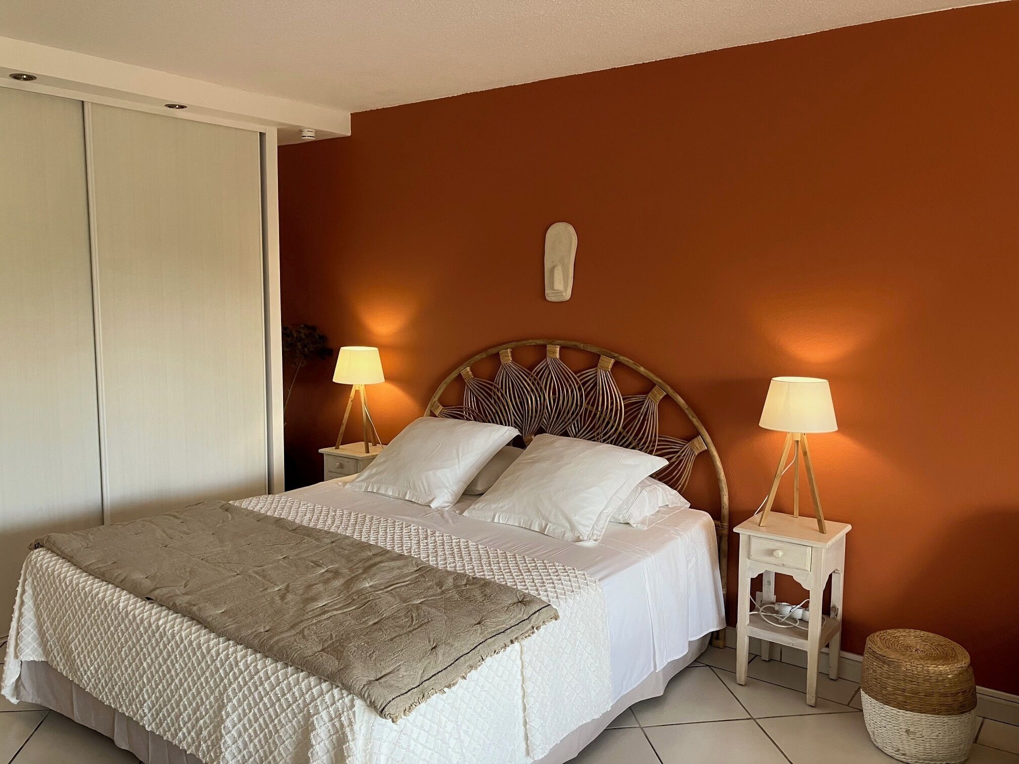 A bedroom with a bed, two bedside tables with lamps, a wall-mounted light fixture, a wardrobe, and a small woven stool.
