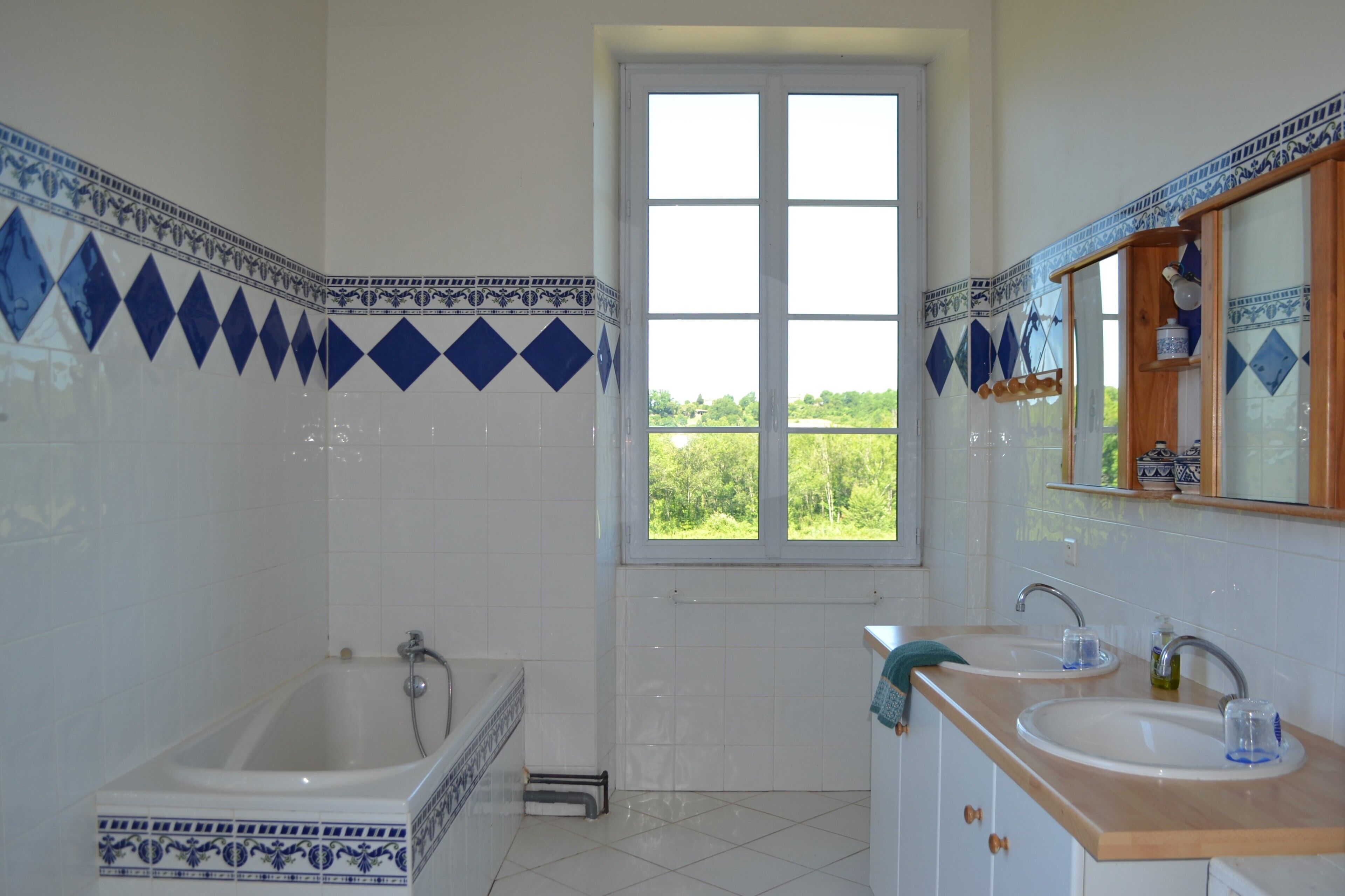 Een badkamer met een badkuip, twee wastafels, een raam met uitzicht en decoratieve blauw-witte tegels.
