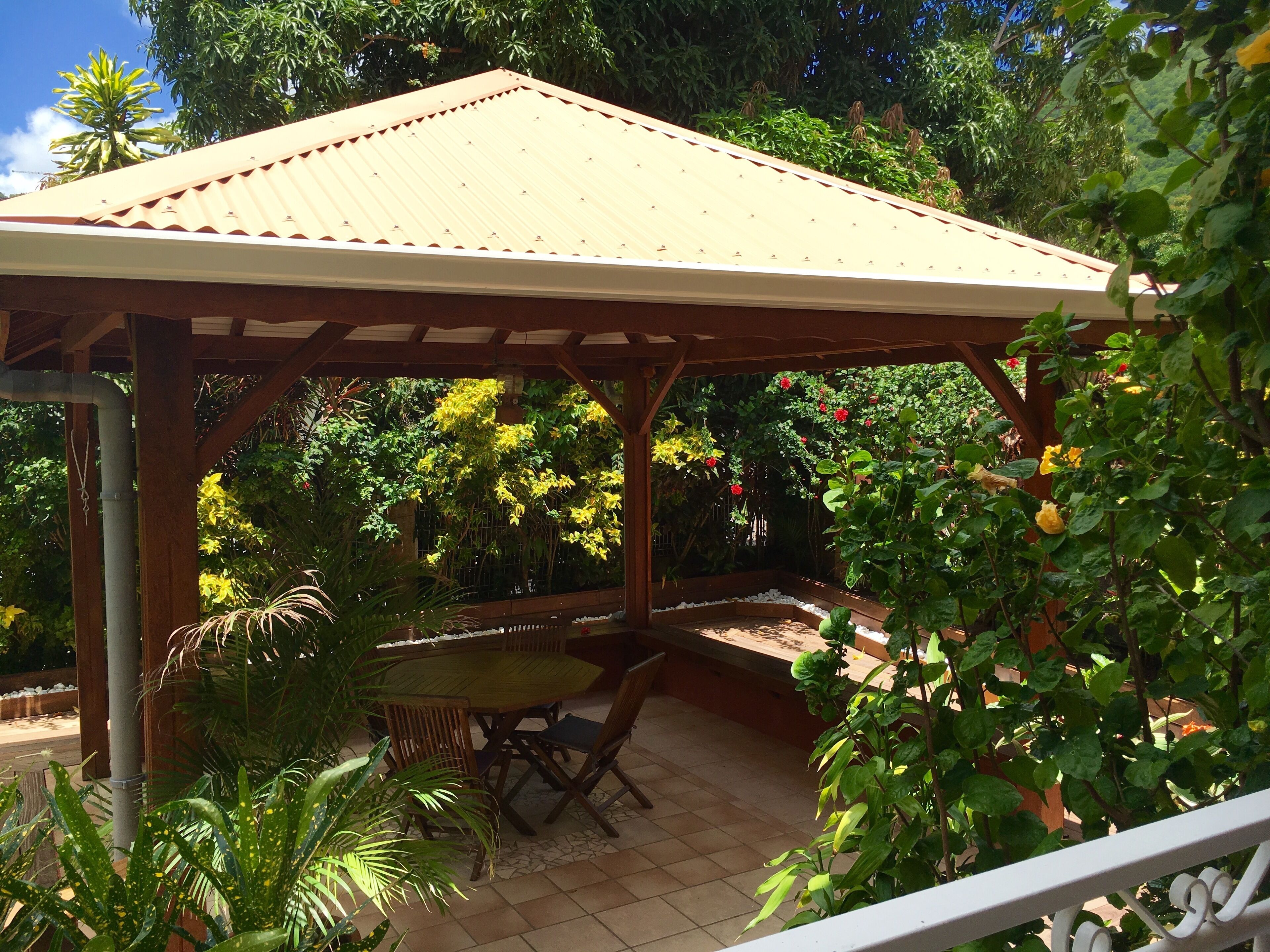 A covered patio area with a table and chairs, surrounded by lush greenery and flowering plants.