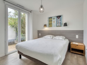 Une chambre à coucher comprenant un lit, une table de chevet, une lampe fixée au mur, un tableau et une porte-fenêtre coulissante donnant sur un espace extérieur.