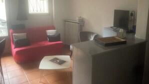 A living room with a red sofa, a small white table, a radiator, a clock on the wall, and a television on a stand.