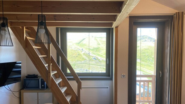 A room with a wooden staircase, a television, a desk, a chair, a window with a view of greenery, and a balcony with a door. - Cozy & Elegant Apartment in Val Thorens, Ski-in/Ski-out, south-facing view, 36m2 (Les Belleville)