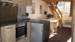 A compact kitchen with wooden cabinets, a stainless steel refrigerator, a built-in oven, and a microwave. There's a small dining area with a table and chairs, a window with a view, and a staircase leading to an upper level. - Cozy & Elegant Apartment in Val Thorens, Ski-in/Ski-out, south-facing view, 36m2 (Les Belleville)