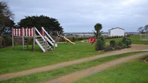 A playground with a slide and climbing structure, a dirt path, and a building in the background. - The terraces of the bay 'between land and sea' (Plounévez-Lochrist)