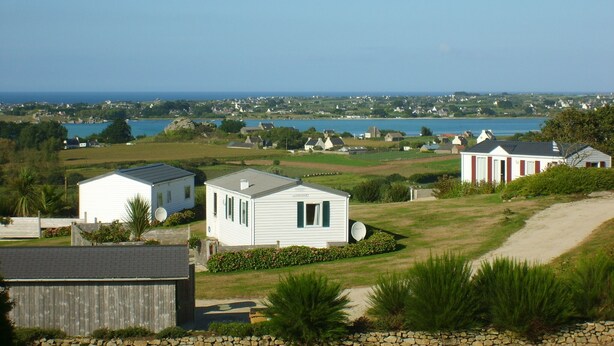 A coastal area with white mobile homes, a stone wall, and a clear view of the sea and distant buildings. - The terraces of the bay 'between land and sea' (Plounévez-Lochrist)