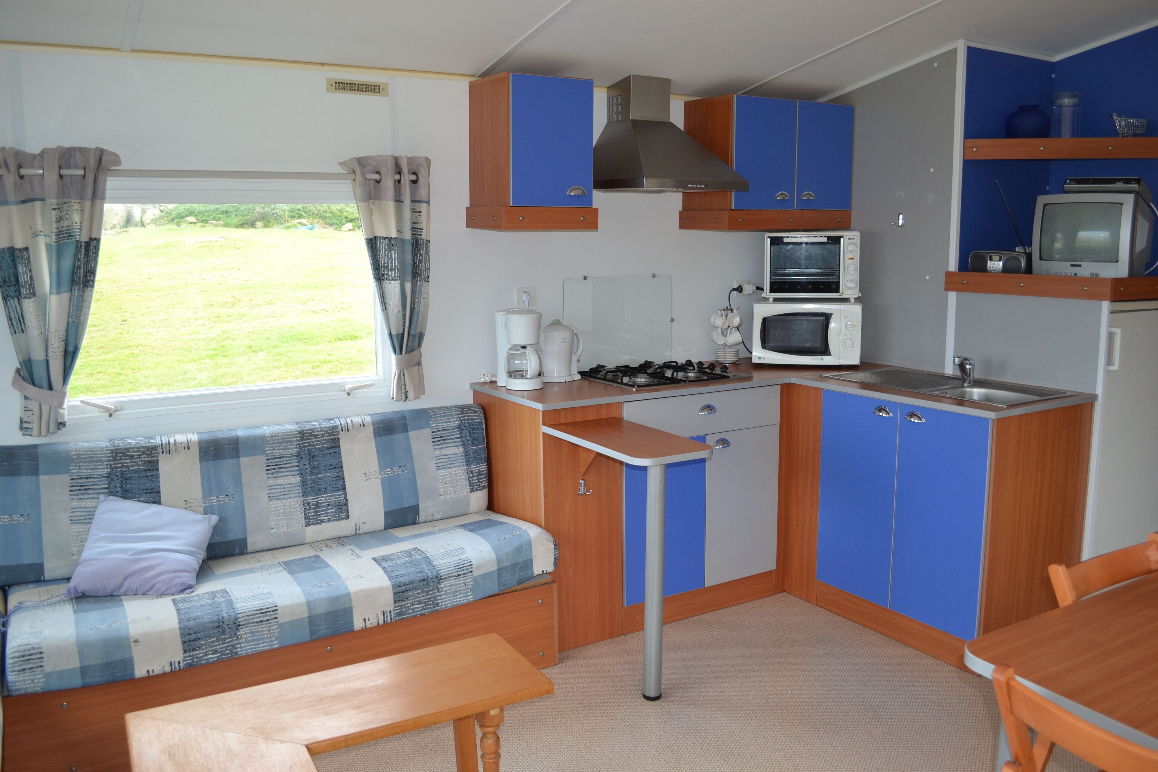 A compact kitchenette with a stove, microwave, and sink. There is a blue and white checkered sofa with a cushion and a small wooden table.