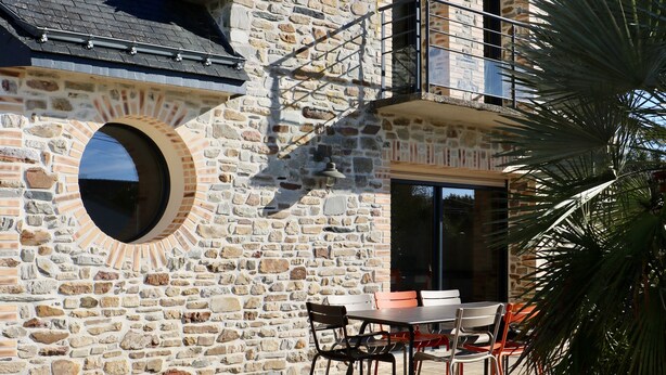 Un bâtiment en pierre doté d’un balcon, d’une fenêtre ronde et d’un espace extérieur aménagé pour s’asseoir.