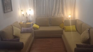 Ein Wohnzimmer mit einem beige farbenen Sofagarnitur, zwei gelben Kissen, einem kleinen runden Tisch mit Lampe und einem gerahmten Bild an der Wand.