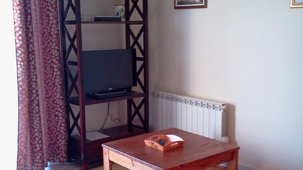 A living room with a TV on a wooden stand, a small wooden table, a sofa, and a radiator on the wall. - Vacation Rental in Cambo-Les-Bains: 2-room, air-conditioned, fully equipped—panoramic view. (Cambo-Les-Bains)